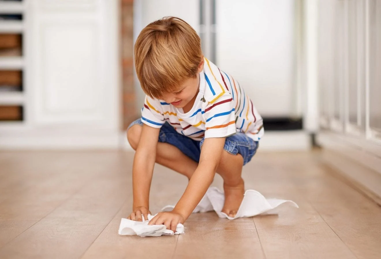 A young boy is crouched down on the floor, carefully wiping up a spill with a cloth. The liquid has spread across the surface, and he looks focused as he cleans, making sure not to miss any spots. The scene shows responsibility and care, as he takes the time to tidy up the mess.