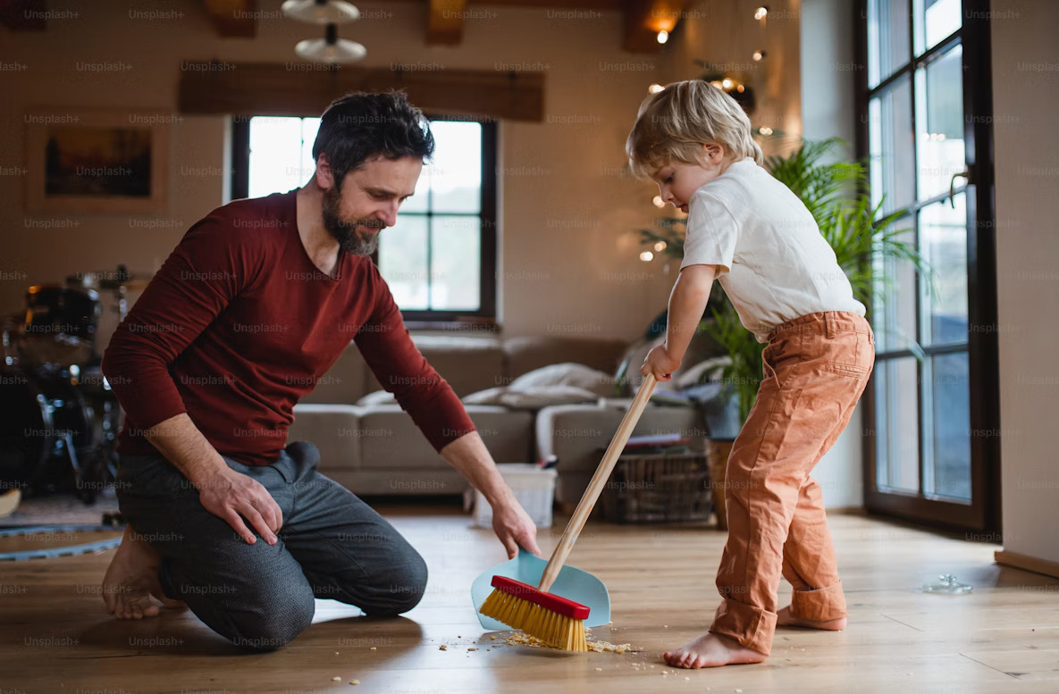A father holds the dustpan steady while his son sweeps dirt in — a simple, hands-on lesson in working together and learning responsibility.